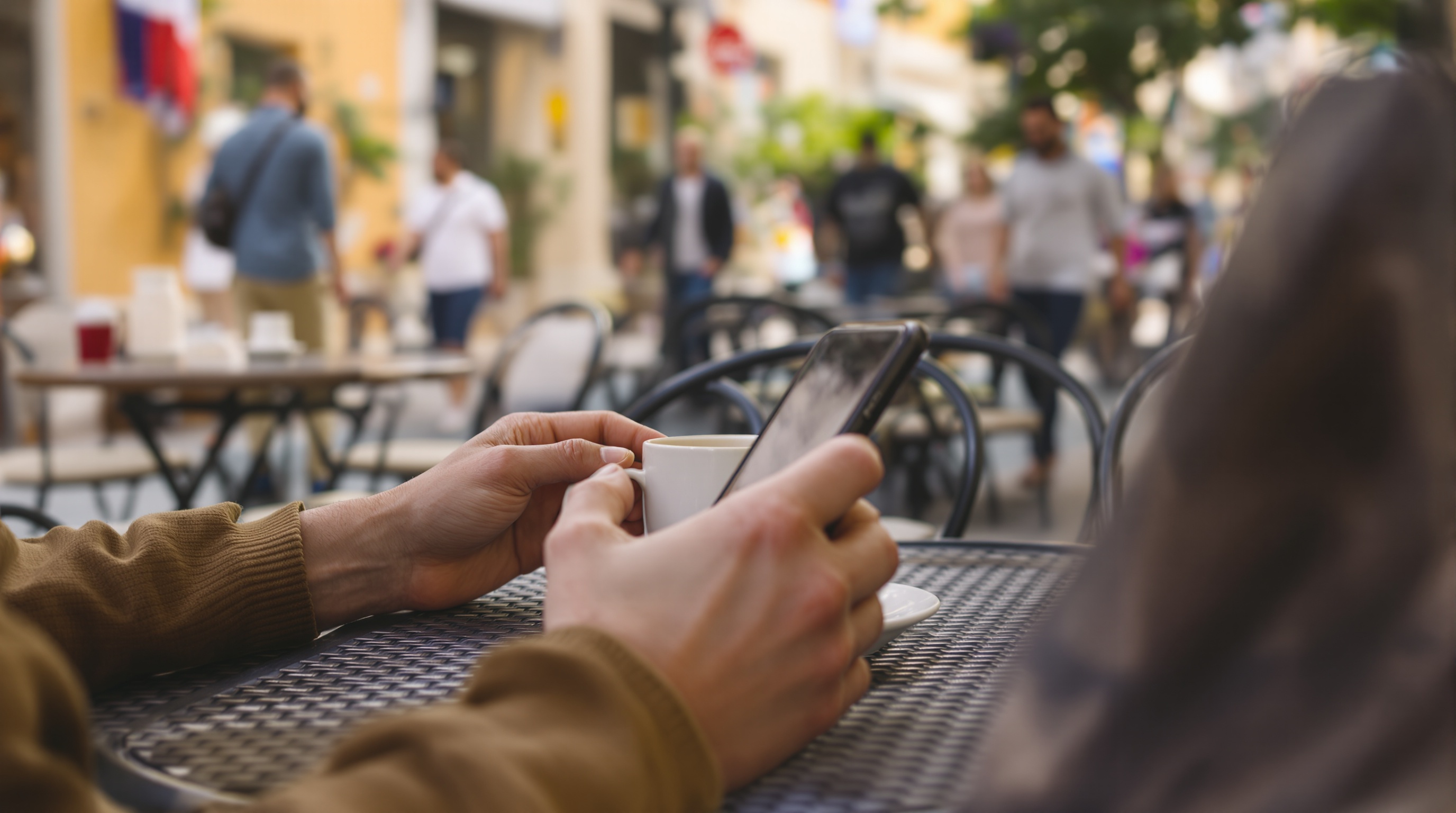 A person holding a phone at a cafe table with their other hand resting on the table, phone screen facing away from camera, natural daylight, candid street photography style