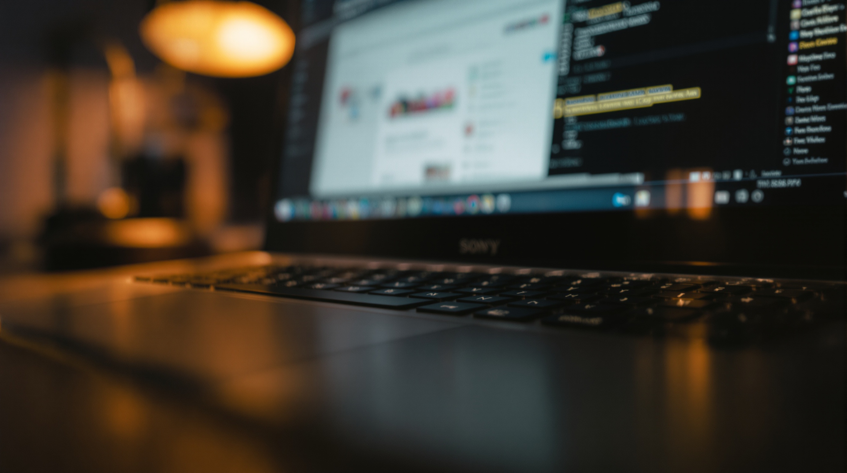 A laptop keyboard in shallow focus with a glowing browser tab reflection visible on the screen, warm desk lamp lighting from the left, muted office tones