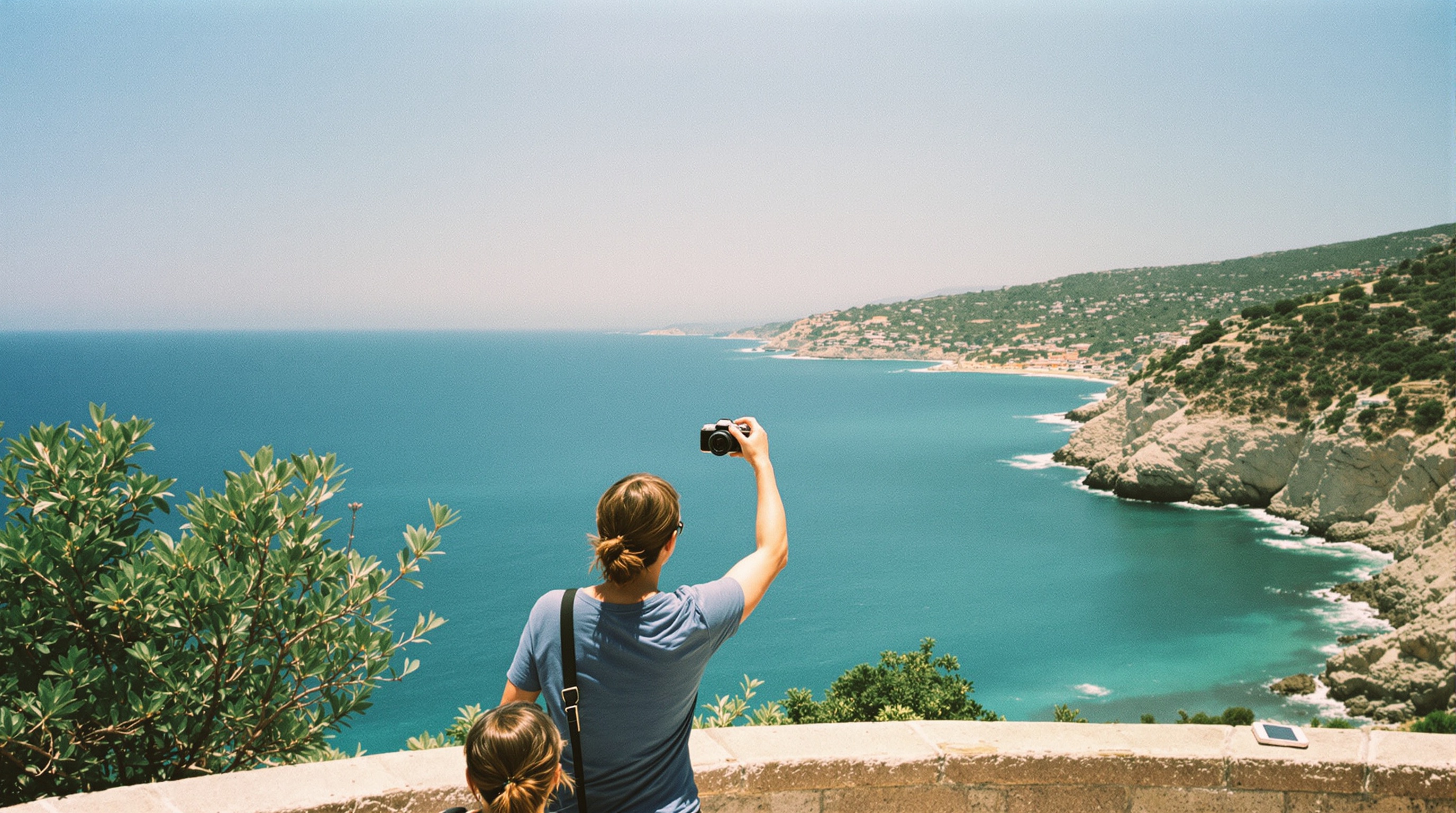 Two people at a Mediterranean coastal viewpoint overlooking turquoise sea, one holding up a camera to share photos with the other