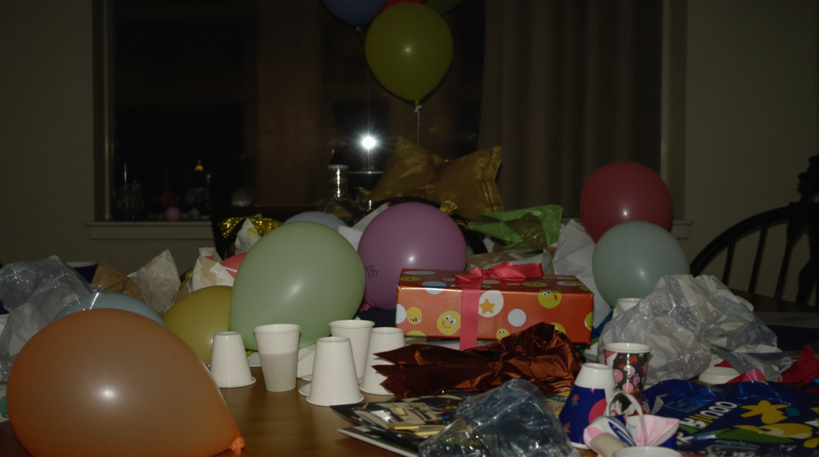 A cluttered table after a birthday party at dusk with deflated balloons, empty cups, and a wrapped gift in soft evening light