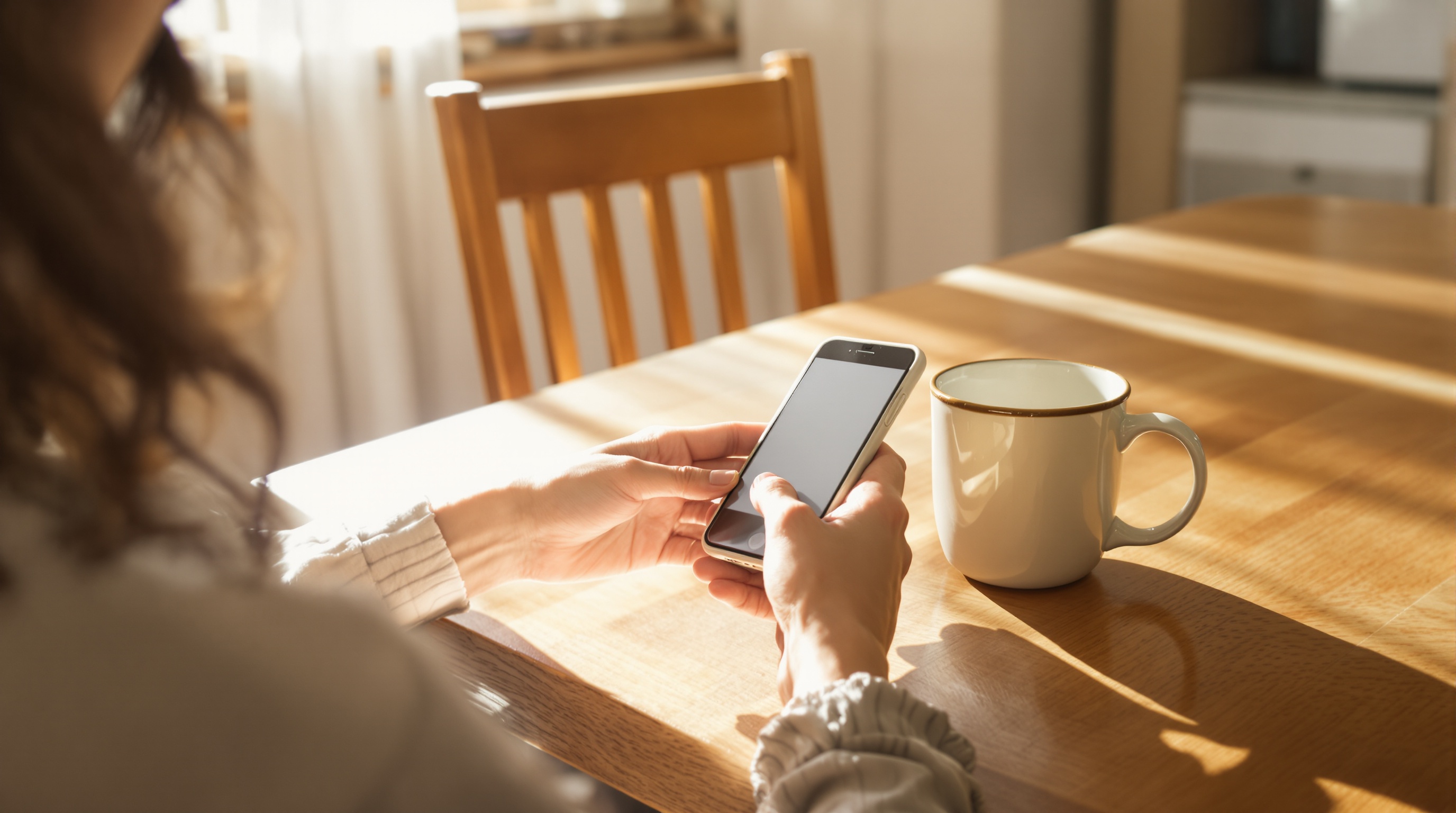 Person sitting at a wooden desk with a cup of coffee, looking at a phone while sunlight streams through a nearby window