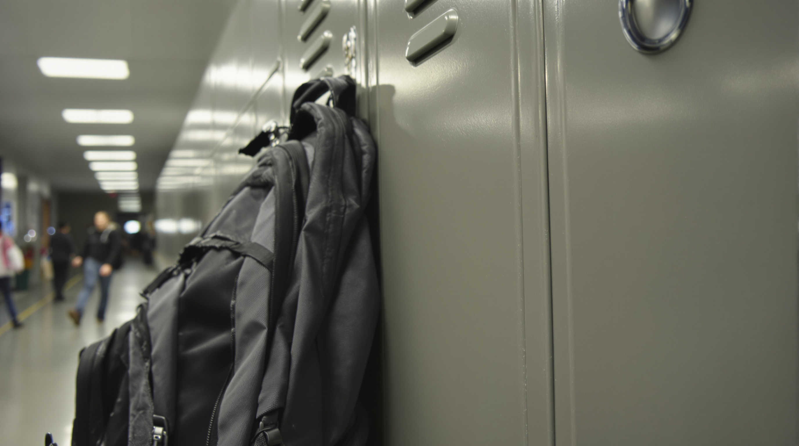 Close-up of a student backpack leaning against a metal locker in a school hallway, combination lock visible, harsh fluorescent overhead lighting, shot on Ricoh GR III, slightly desaturated tones, ISO 800 grain
