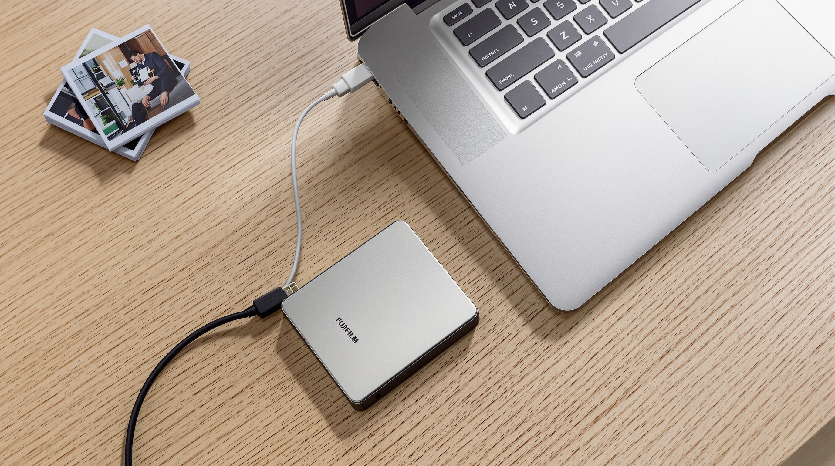 External hard drive connected to a laptop on a wooden desk, with a stack of photo prints and a USB cable nearby