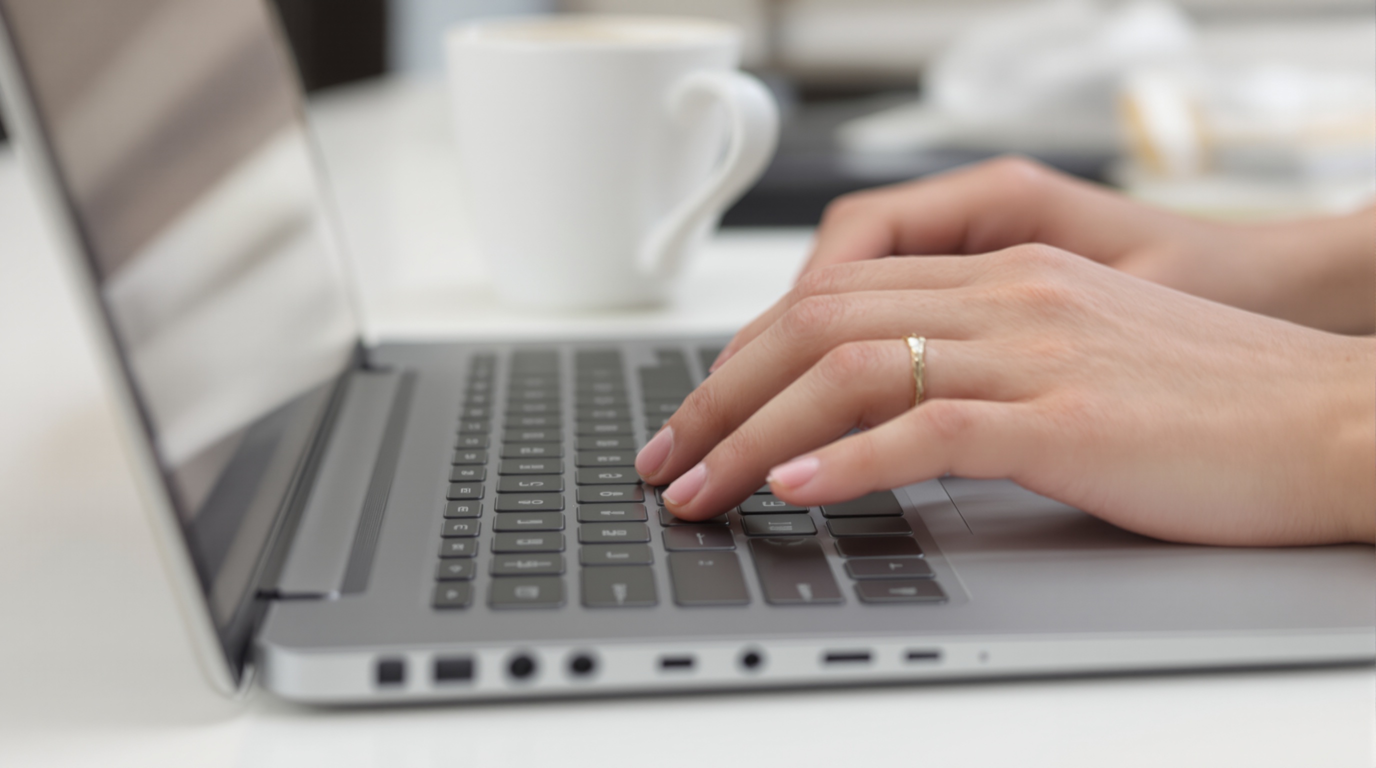 Close-up of hands typing on a laptop keyboard seen from the side, coffee cup nearby, soft office lighting