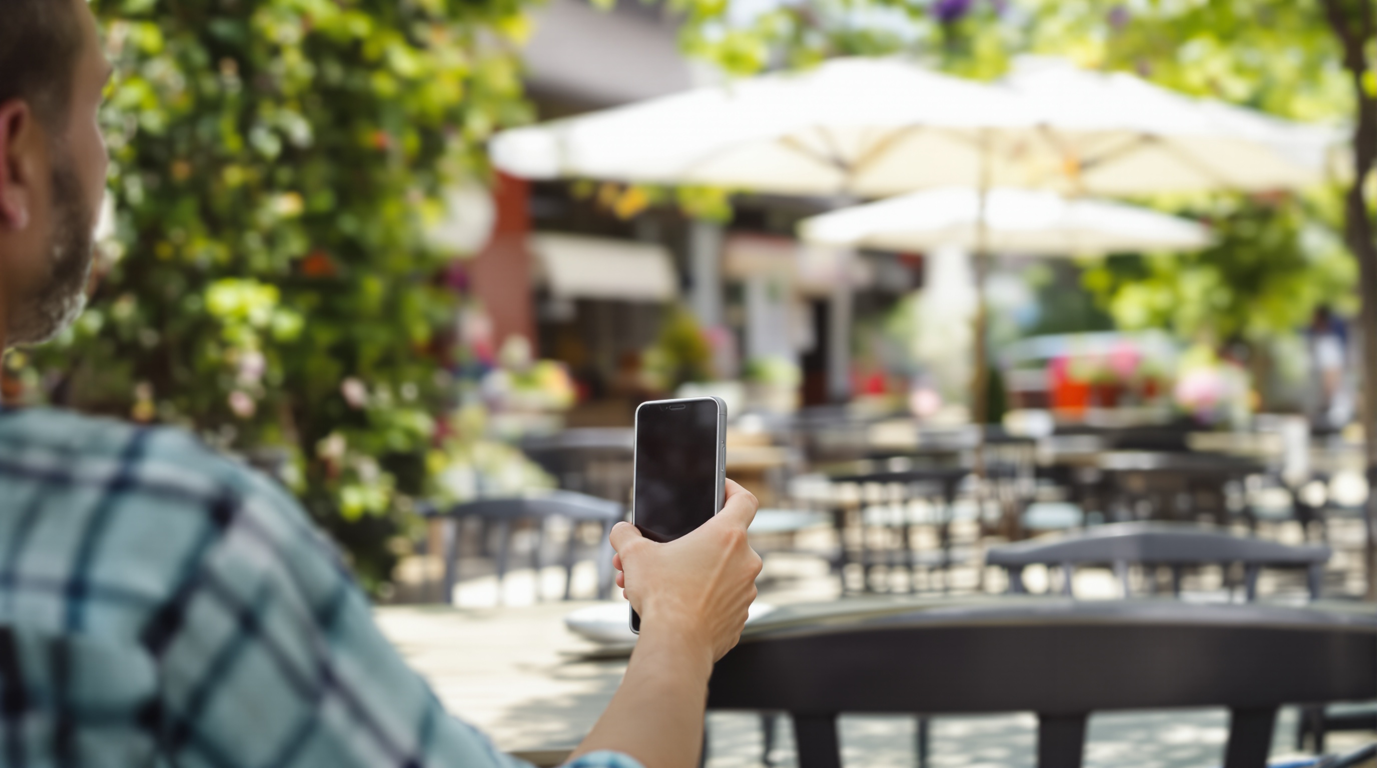 A person holding a smartphone while sitting at a cafe table, photographed from behind showing the phone screen from a distance