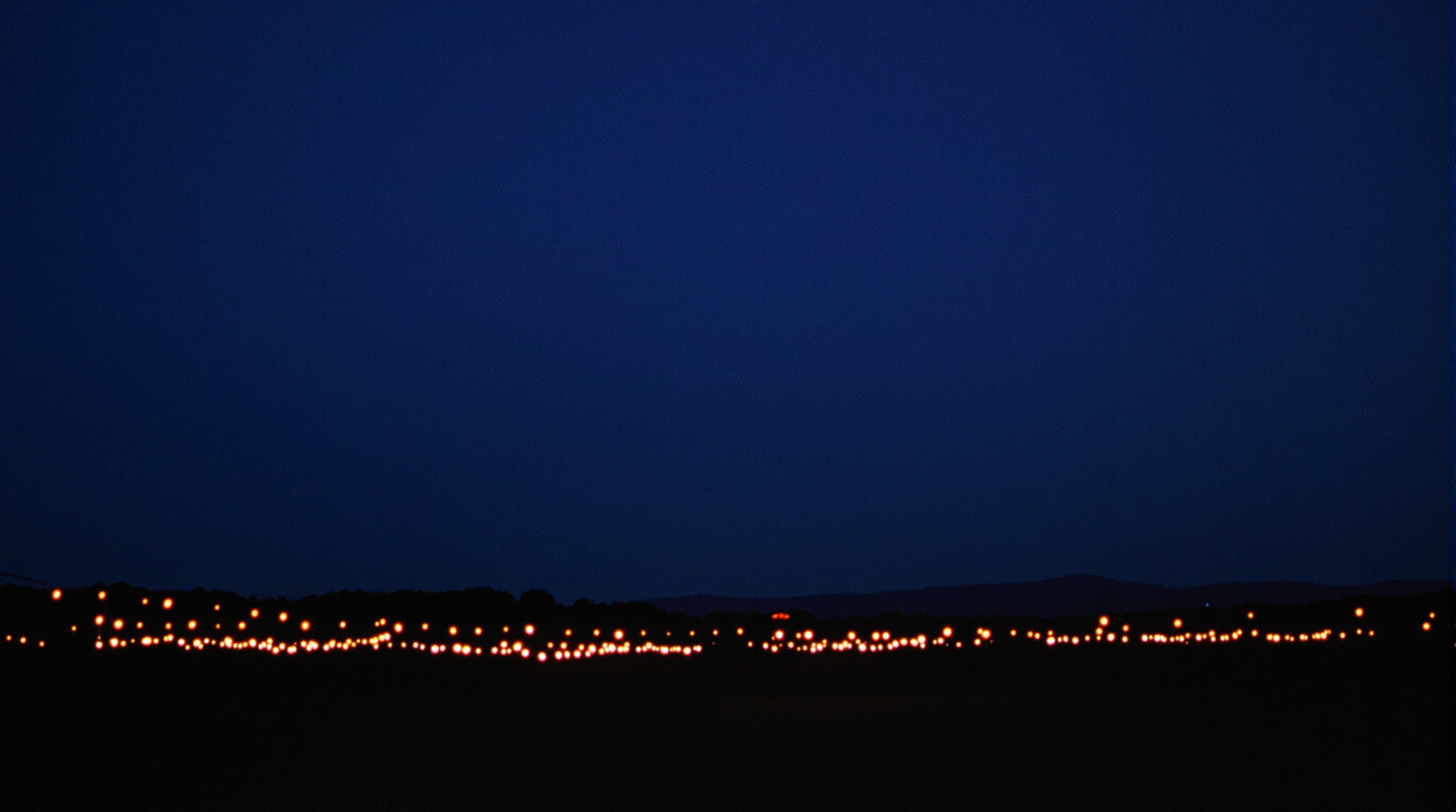 A festival field at dusk with string lights and a distant stage glow as a few people walk away, the weekend that lives on in one shared album