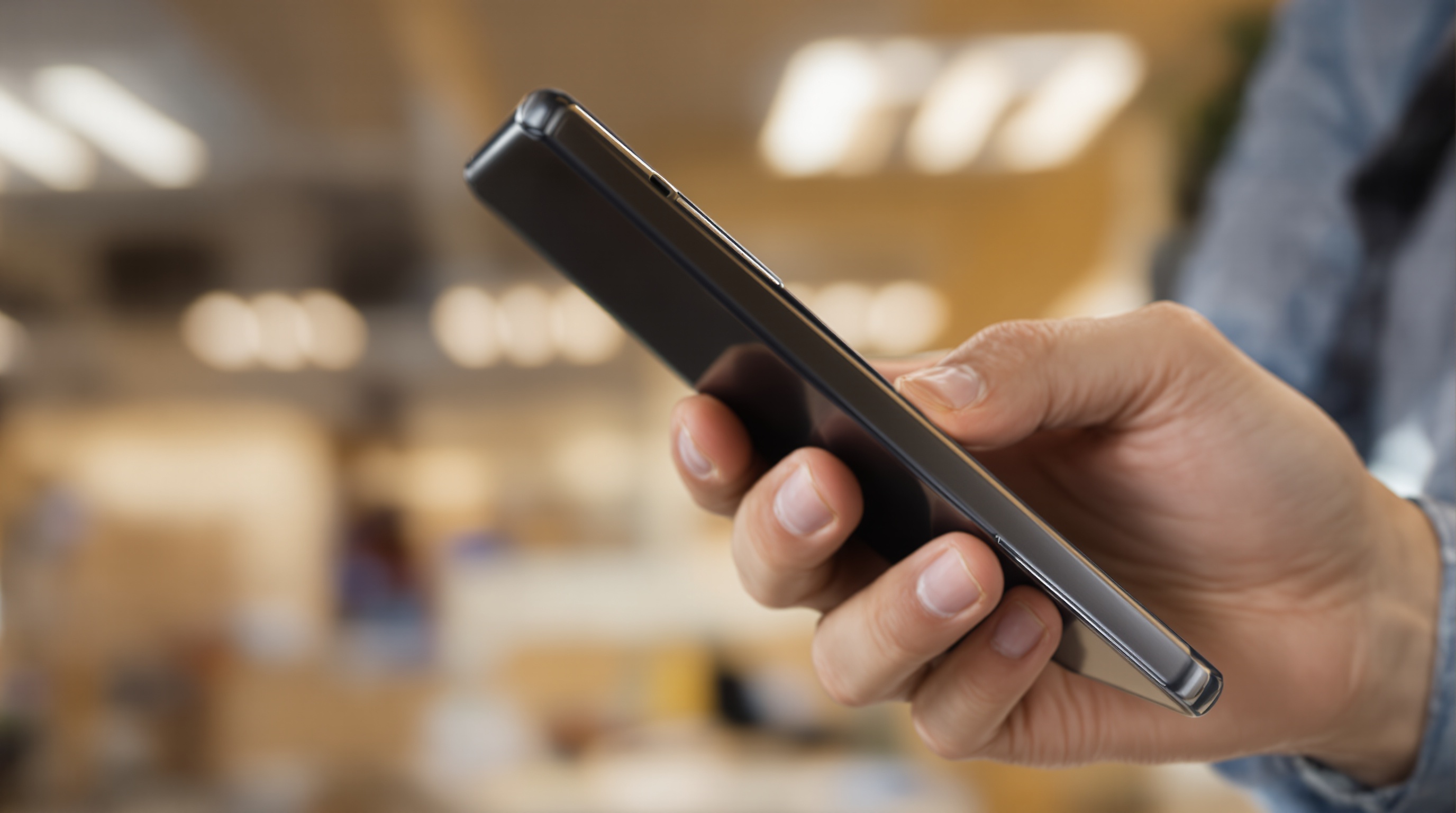 Close-up of fingers holding a Samsung phone from the side, out-of-focus office background with warm overhead lighting
