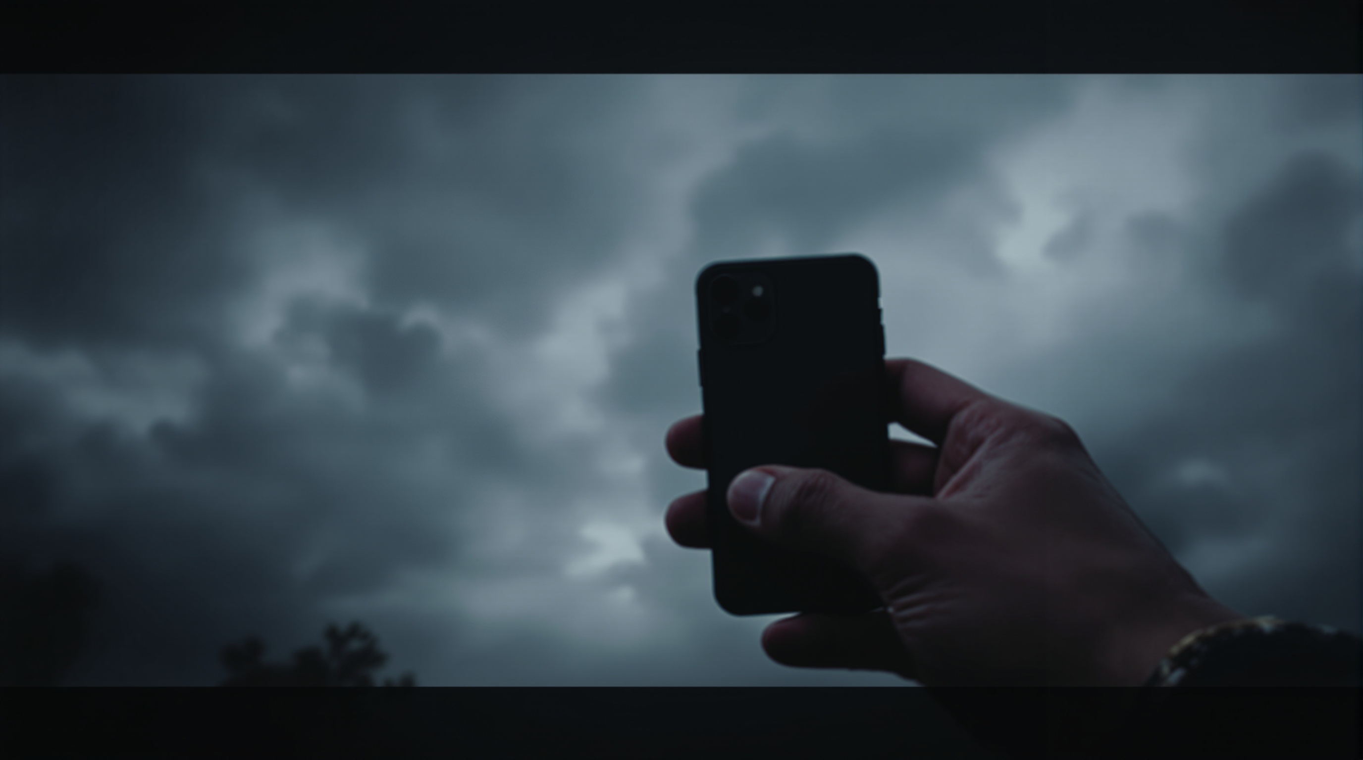 Close-up of a hand holding an iPhone with camera app visible from behind at a low angle against cloudy sky