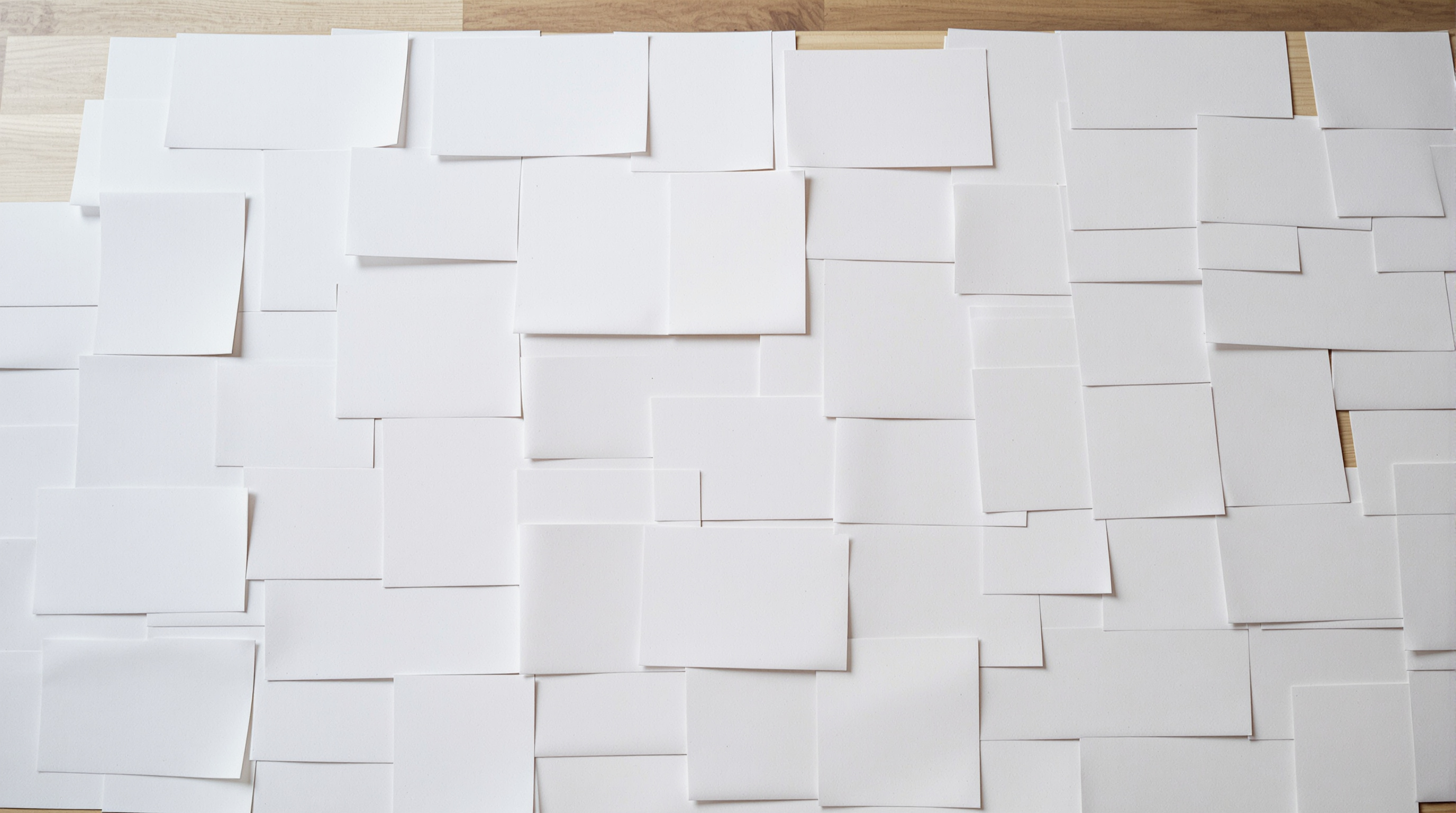 Hundreds of blank white photo prints laid in an overlapping layer across a pale oak table, seen from directly above in soft daylight