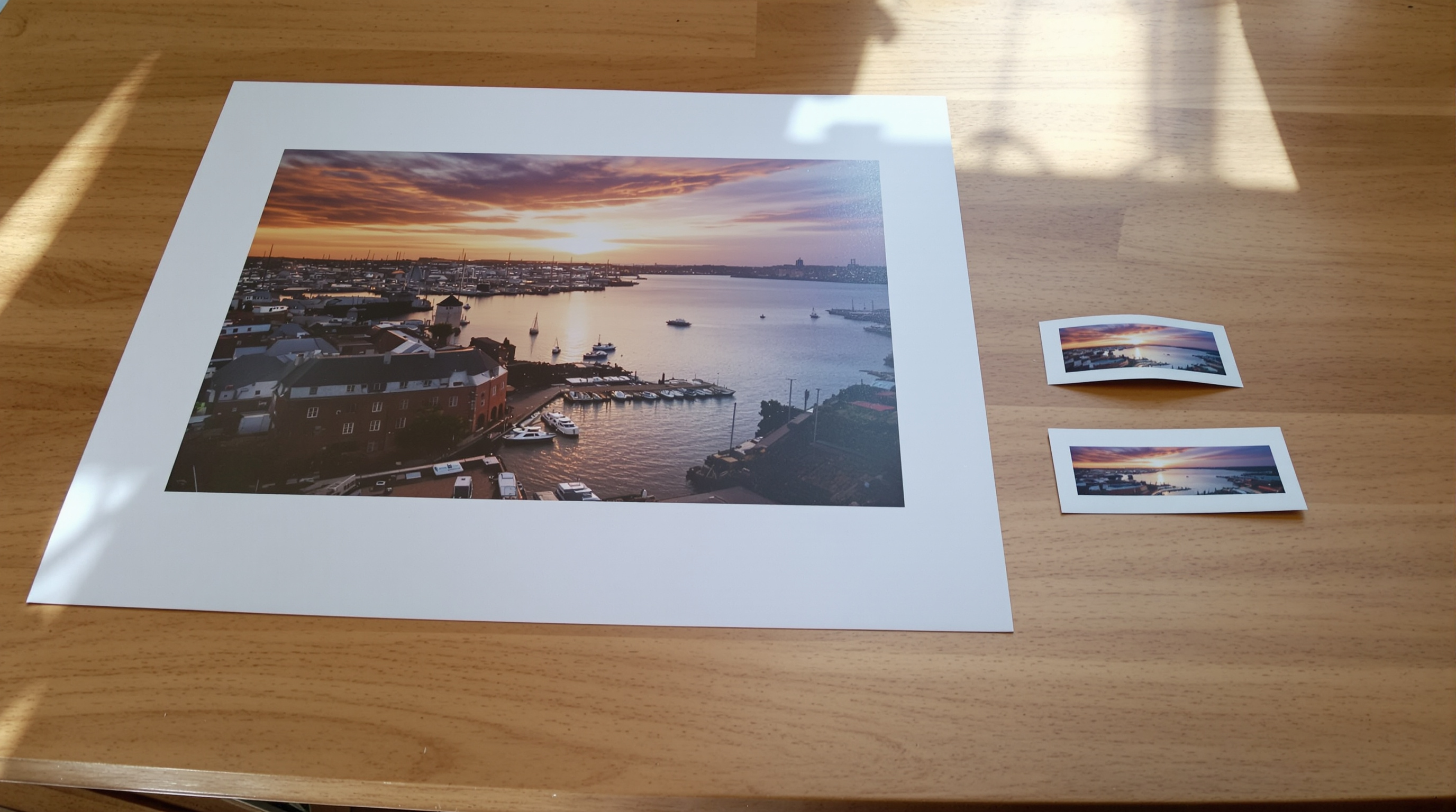 A large print of a harbour at sunrise on an oak table beside two much smaller prints of the identical scene