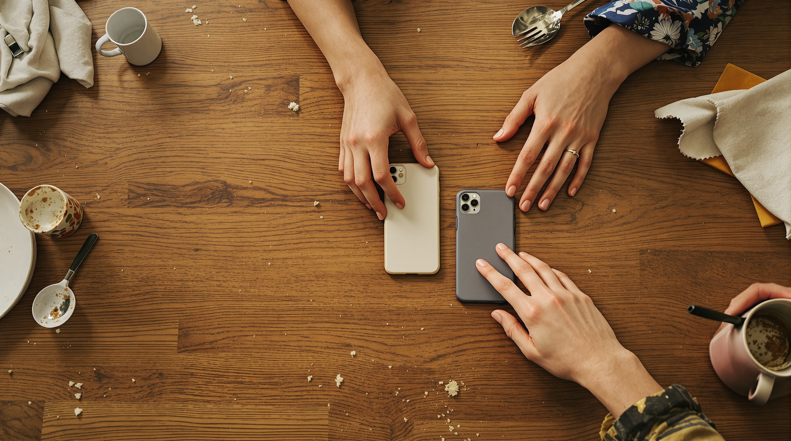 Two hands passing a phone across a wooden dining table, both phones different sizes and cases, remains of a shared meal around them