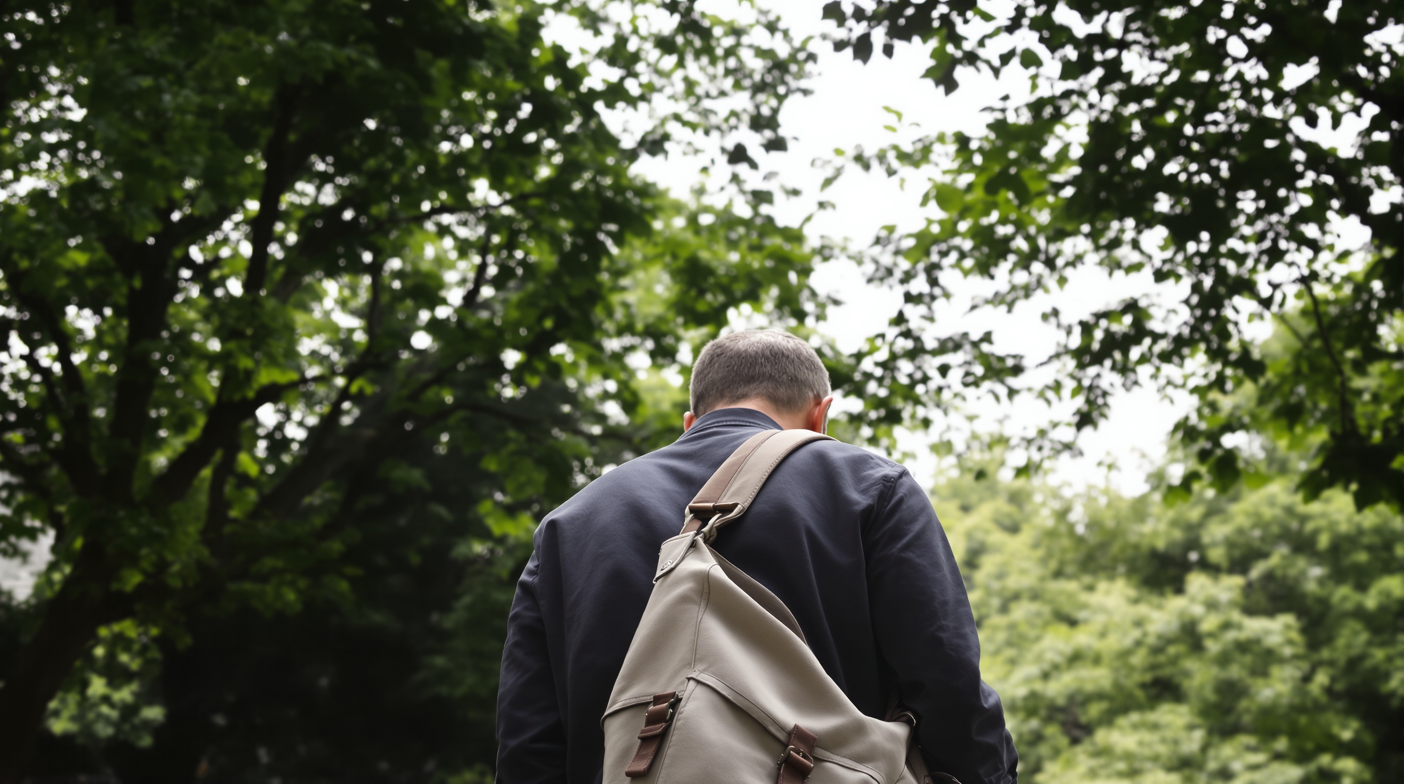 A person walking through a park, photographed from behind, with a camera bag over their shoulder