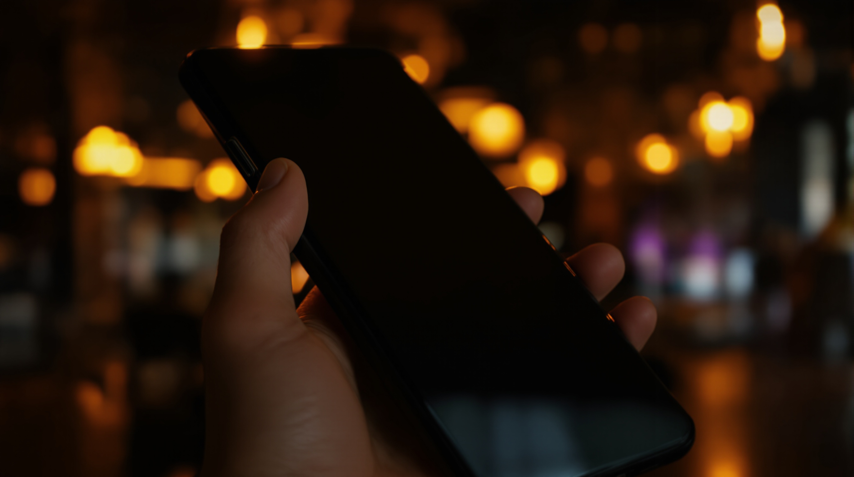 Close-up of a hand holding a phone with the screen turned away, in a dimly lit bar setting