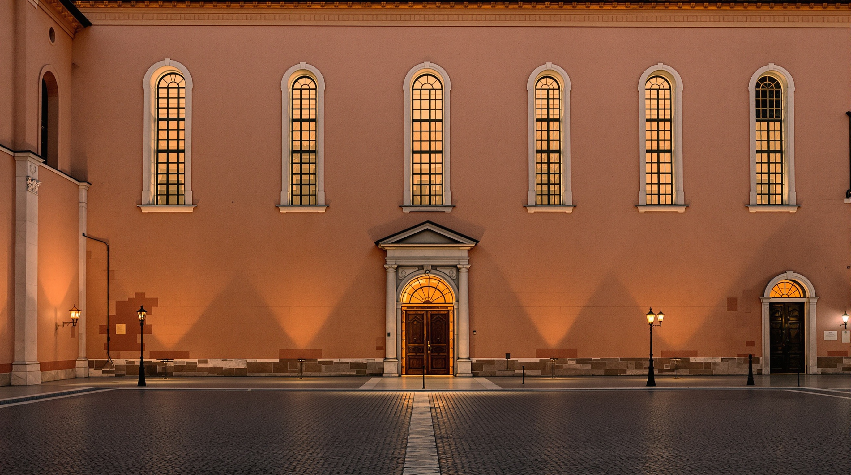 Classical European museum facade at dusk with warm stone walls and tall arched windows, long shadows across the courtyard